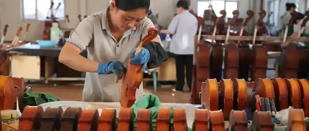 A luthier carefully gluing a fingerboard to a violin neck on a workbench surrounded by tools.